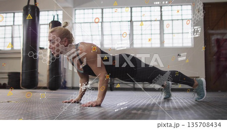 Female athlete wearing sportswear performing plank push-up on gym mat, with punching bags Female athlete wearing sportswear performing plank push-up on gym mat, with punching bags 135708434