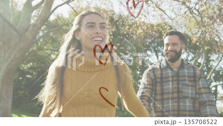 Walking couple in mustard sweater and plaid shirt holding hands in autumn park, with heart overlays 135708522