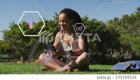 Smiling woman holding smartphone and wearing smartwatch while sitting on park lawn, with backpack Smiling woman holding smartphone and wearing smartwatch while sitting on park lawn, with backpack 135708528