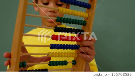 Boy wearing yellow shirt gripping abacus while focusing on colorful beads against green chalkboard 135708589