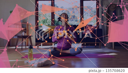 Meditating woman wearing purple top and leggings on yoga mat in living room, with water bottle Meditating woman wearing purple top and leggings on yoga mat in living room, with water bottle 135708620