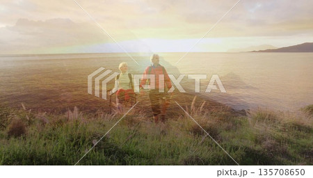 Walking couple of hikers holding poles and backpacks on grassy coastal bluff at sunset, warm glow 135708650