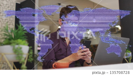 Man wearing burgundy shirt scrolling smartphone in office with laptop plant mug and map overlay Man wearing burgundy shirt scrolling smartphone in office with laptop plant mug and map overlay 135708728