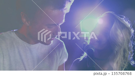 Singing duet partners wearing white dark outfits leaning toward mic on club stage, with lights haze 135708768