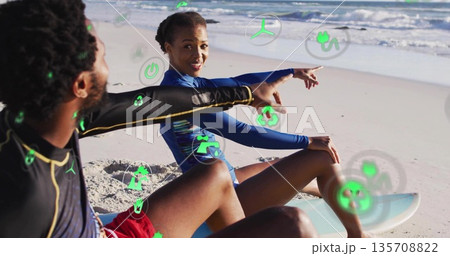 Two surfers in rash guards pointing at waves astride surfboards at shore, with green digital icons Two surfers in rash guards pointing at waves astride surfboards at shore, with green digital icons 135708822