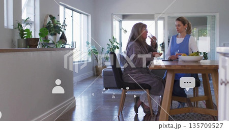 Senior woman eating yogurt at home table, caregiver in apron holding glass with fruit bowl, plants 135709527