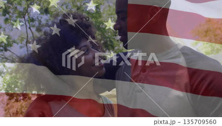 Couple wearing pink tee, grey raglan exchanging smiles in sunlit park, translucent US flag overlay 135709560