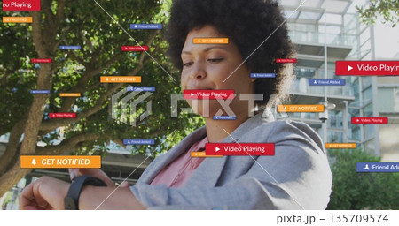 Glancing woman in blazer checking smartwatch with floating notifications in plaza, copy space 135709574