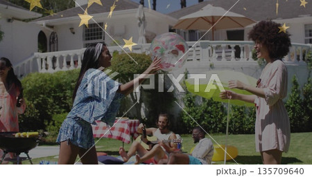 Playing friends tossing clear beach ball with fruit-motif on sunny backyard lawn, picnic gathering 135709640