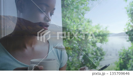 Standing woman wearing teal top holding white mug and smartphone by window viewing lake, copy space 135709646