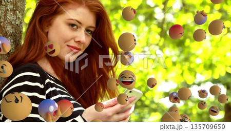 Redhead woman leaning against trunk holding smartphone gazing at camera in park, with emoji spheres Redhead woman leaning against trunk holding smartphone gazing at camera in park, with emoji spheres 135709650