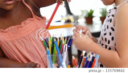 Two girls wearing peach-pink top and spotted dress grabbing from container at desk, colored pencils Two girls wearing peach-pink top and spotted dress grabbing from container at desk, colored pencils 135709682