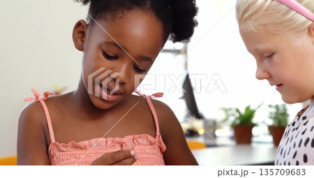 Inspecting two friends holding small craft piece at bright play area table, with potted plants Inspecting two friends holding small craft piece at bright play area table, with potted plants 135709683