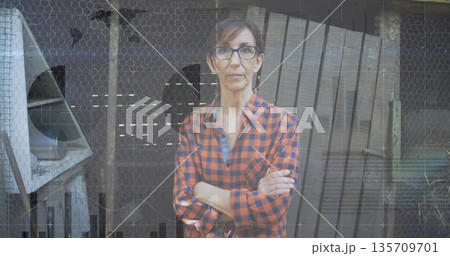 Standing mature woman crossing arms in front of barn wire mesh barrier, with metal fan housing Standing mature woman crossing arms in front of barn wire mesh barrier, with metal fan housing 135709701