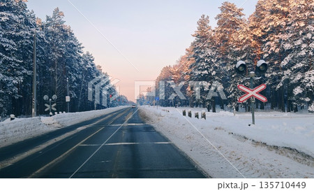 Winter Road with Railroad Crossing Sign in Snowy Pine Forest. Snow-covered rural road with railroad crossing barrier and lights, car driving through pine forest at sunset, winter countryside scene. 135710449