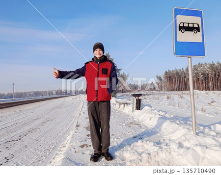 Young Man Hitchhiking at Winter Bus Stop Sign. Young man in warm jacket giving thumbs up at snowy rural bus stop sign with bus icon, winter road in pine forest background. Outdoor seasonal scene. 135710490