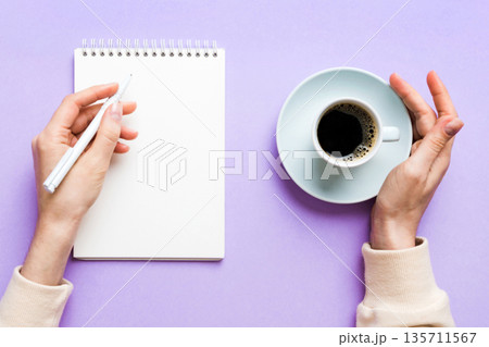 Woman hand with pencil writing on notebook and hold coffee cup. Woman working on office table with coffee Woman hand with pencil writing on notebook and hold coffee cup. Woman working on office table with coffee 135711567