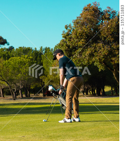 Man leaning over golf ball beside golf bag on green and lining up putt with putter Man leaning over golf ball beside golf bag on green and lining up putt with putter 135711883