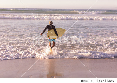 African American man holding yellow surfboard with black rash guard and walking into waves at beach African American man holding yellow surfboard with black rash guard and walking into waves at beach 135711885