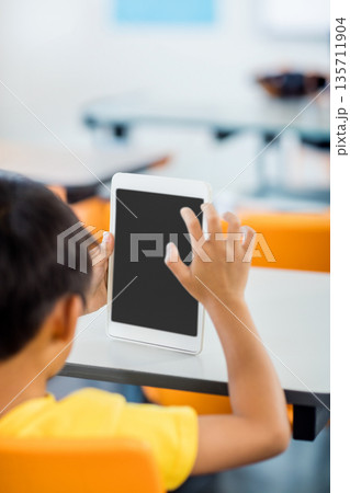 White tablet being held upright on white desk with blank screen in classroom, with orange chairs White tablet being held upright on white desk with blank screen in classroom, with orange chairs 135711904