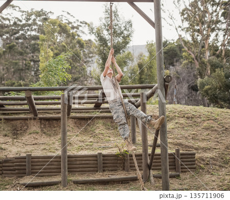 Male military trainee wearing camo climbing rope on wooden frame obstacle course in grassy clearing Male military trainee wearing camo climbing rope on wooden frame obstacle course in grassy clearing 135711906