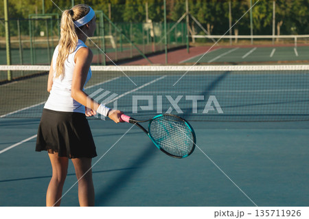 Female athlete wearing tennis attire, gripping tennis racket and facing net on outdoor blue court Female athlete wearing tennis attire, gripping tennis racket and facing net on outdoor blue court 135711926