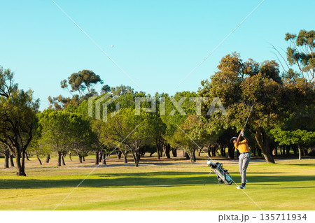 African American male golfer swinging club sending golf ball on fairway beside golf bag, copy space African American male golfer swinging club sending golf ball on fairway beside golf bag, copy space 135711934