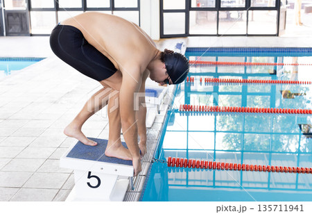 Male athlete crouching on starting block at lap pool wearing swim cap and goggles, copy space 135711941