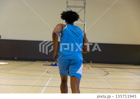 African American male basketball player running toward hoop at gym near orange cones, water bottle African American male basketball player running toward hoop at gym near orange cones, water bottle 135711945