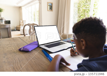 African American boy writing in notebook with pencil at table in room with laptop, copy space 135711946
