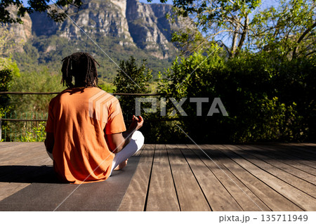 African American man meditating on yoga mat on wooden deck by metal railing wearing orange t-shirt African American man meditating on yoga mat on wooden deck by metal railing wearing orange t-shirt 135711949