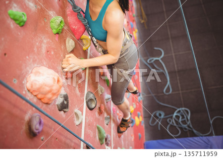 Woman climbing red wall in gym gripping holds, wearing harness with belay rope, climbing shoes 135711959