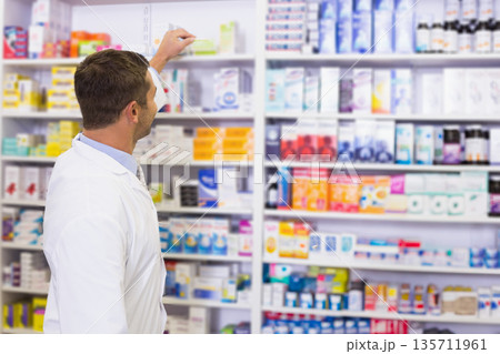 Male pharmacist wearing lab coat selecting medication from pharmacy shelving at counter, copy space 135711961