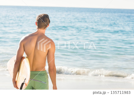 Shirtless man with green boardshorts holding longboard on shore gazing at turquoise sea, copy space 135711983