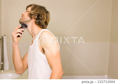 Man wearing white tank top standing at bathroom counter grooming beard with electric razor Man wearing white tank top standing at bathroom counter grooming beard with electric razor 135712004