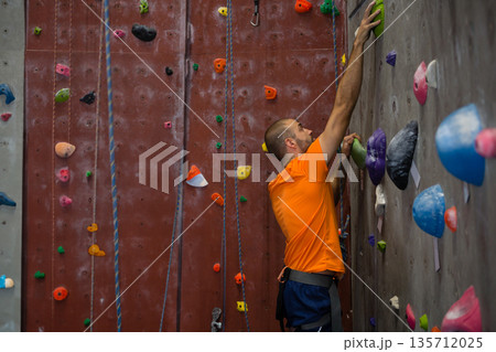 Male climber ascending gym wall gripping green hold on grey panel wearing blue harness, copy space Male climber ascending gym wall gripping green hold on grey panel wearing blue harness, copy space 135712025