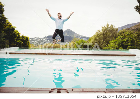 Man wearing blue shirt leaping off wooden deck beside turquoise pool raising arms toward peak Man wearing blue shirt leaping off wooden deck beside turquoise pool raising arms toward peak 135712054