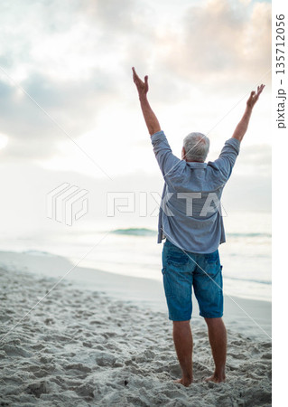 Senior man standing barefoot on sandy shoreline facing ocean raising arms toward sky with waves 135712056