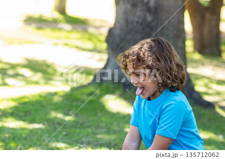 Curly-haired boy sticking out tongue while kneeling on grassy lawn in sunlit park, copy space Curly-haired boy sticking out tongue while kneeling on grassy lawn in sunlit park, copy space 135712062
