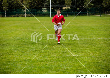 Male rugby player wearing red jersey sprinting while holding white ball on pitch before goalposts 135712071