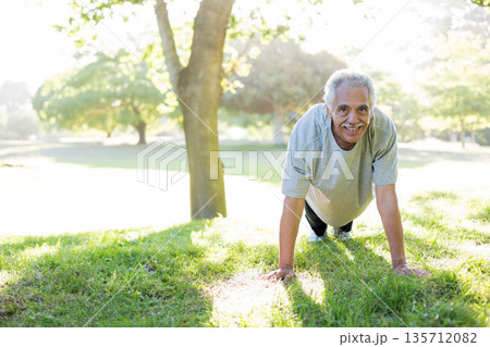 Senior man doing push-ups by tree trunk in park wearing sports shirt, pants, sneakers copy space 135712082