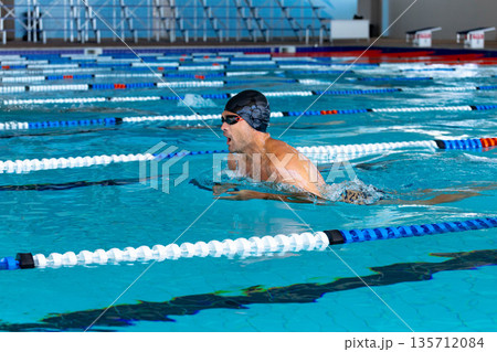 Male swimmer wearing cap and goggles gliding in breaststroke across lap pool toward starting blocks 135712084