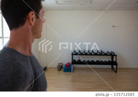 Man in grey t-shirt picking kettlebell from two-tier rack beside yoga mats in gym, copy space 135712085
