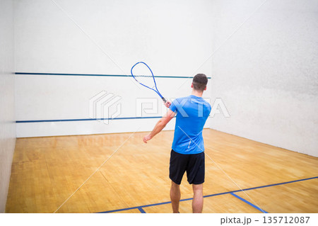 Squash player swinging racket on wooden floor, aiming shot against white wall marked by blue lines 135712087
