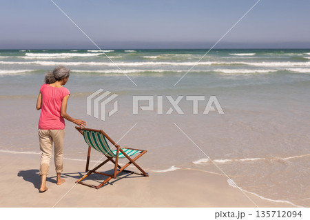 Senior African American woman positioning striped beach chair on wet sand at ocean edge, copy space 135712094