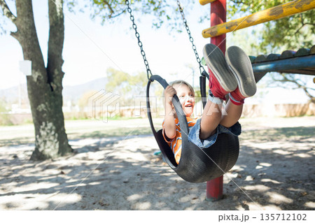 Boy leaning back on swing seat hanging from chains on red post in playground, copy space Boy leaning back on swing seat hanging from chains on red post in playground, copy space 135712102