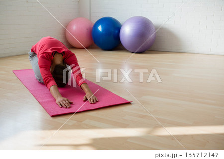 Woman wearing red top performing child's pose on pink yoga mat in gym with fitness balls Woman wearing red top performing child's pose on pink yoga mat in gym with fitness balls 135712147