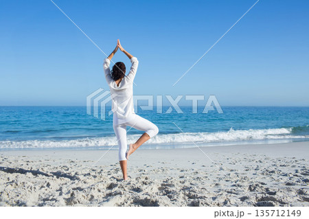 Woman practicing yoga in sportswear on sandy beach at water's edge with ocean waves and horizon 135712149