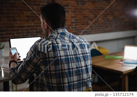 Man using smartphone and blank-screen laptop at wooden desk with potted plant, copy space 135712174