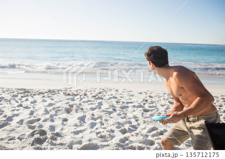 Shirtless male standing on beach by turquoise sea, preparing to throw light blue disc, copy space 135712175